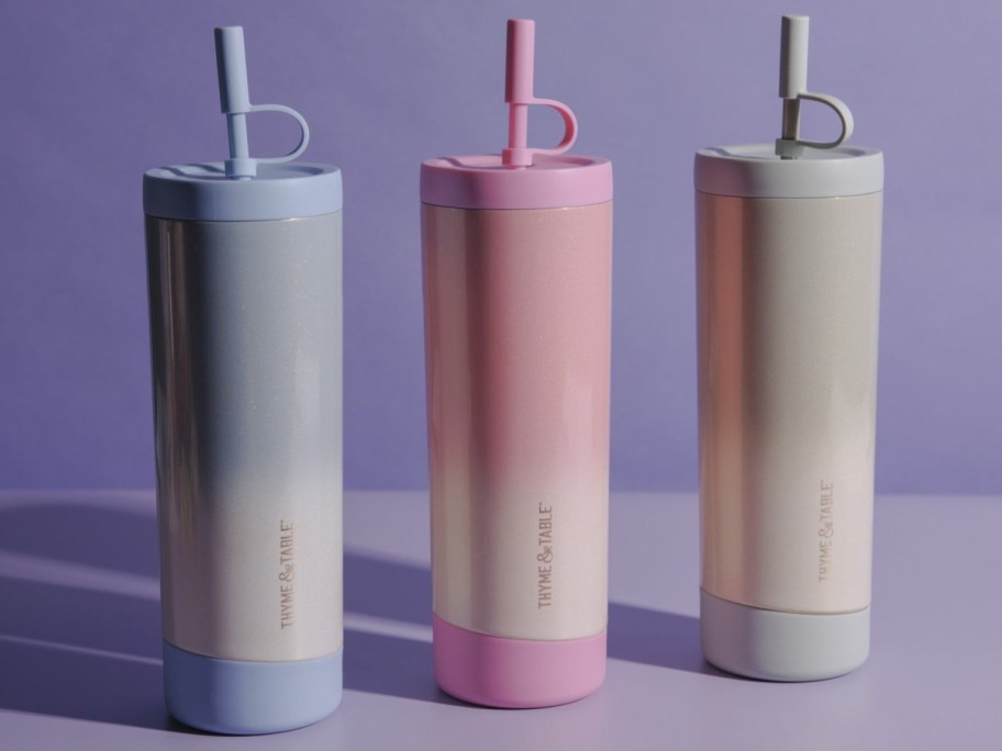 Three stylish, gradient-colored tumblers in blue, pink, and gray with lids and straws, placed on a purple background.