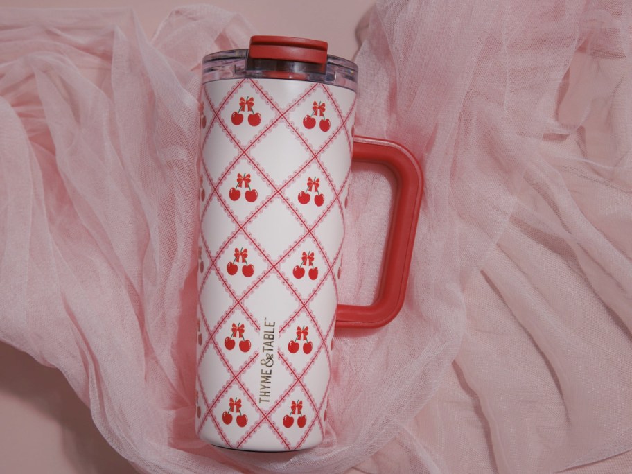 A decorative travel mug with a red handle and a white body featuring a diamond pattern adorned with cherries, resting on a soft, pink fabric background.