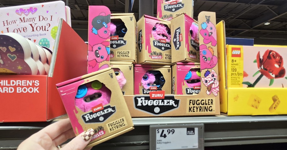 hand holding up boxed Zuru Baby Fuggler keychain in front of stack of more boxes