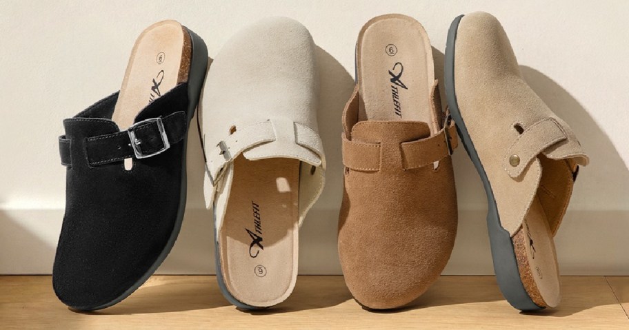 Four suede clogs in black, cream, brown, and beige are lined up on a wooden surface. Each has a decorative buckle.