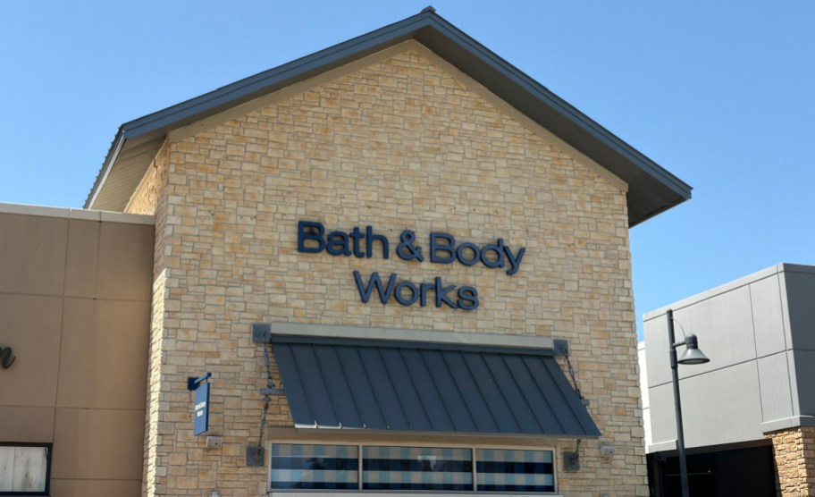 The exterior storefront of a Bath & Body Works store featuring a light tan stone facade, blue signage, and a dark metal awning under a clear blue sky.