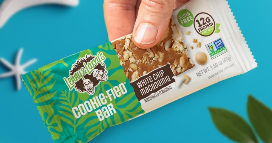 A hand holds a Lenny & Larry's Cookie-fied Bar in colorful green packaging with white chip macadamia flavor. Icons indicate it's gluten-free, non-GMO, and contains 12g of protein. The background is bright blue with a hint of green leaves, conveying a fresh and appealing tone.