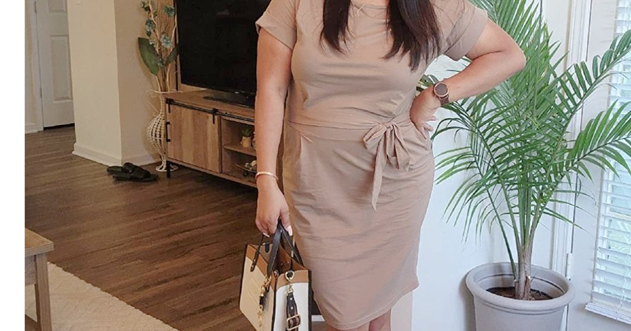 A woman in a beige dress, standing confidently in a living room with wooden floors. She holds a white handbag and is near a potted plant by a window.