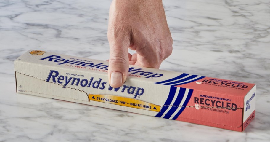 A hand is about to open a box of Reynolds Wrap recycled aluminum foil on a marble countertop. The box is predominantly white with blue and red accents.