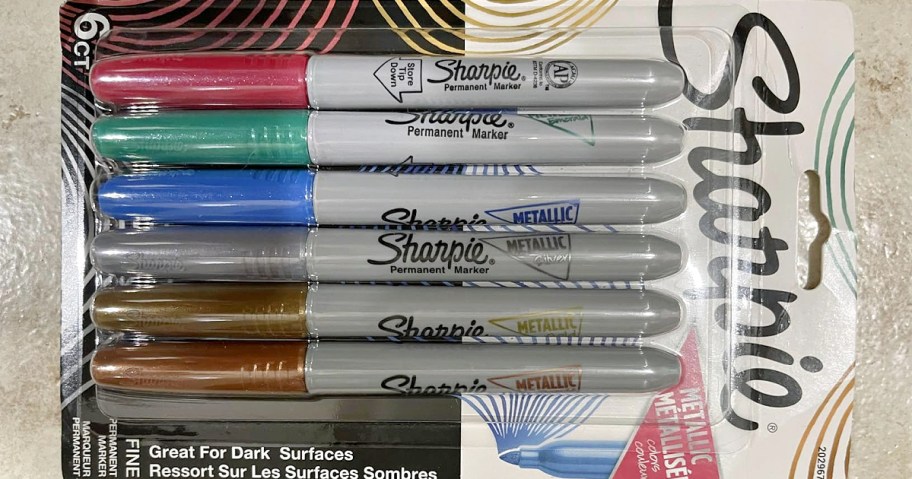 Six Sharpie metallic permanent markers in various colors (pink, green, blue, silver, gold, copper) are in packaging labeled "Great for Dark Surfaces."