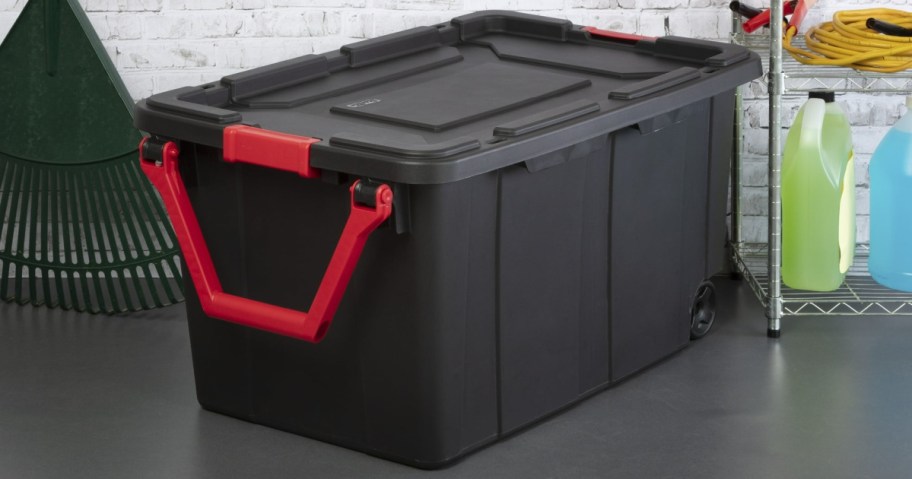 Large black storage bin with red handles sits on a gray floor. Nearby are a green rake, shelves with colorful containers, a coiled hose, and a brick wall.