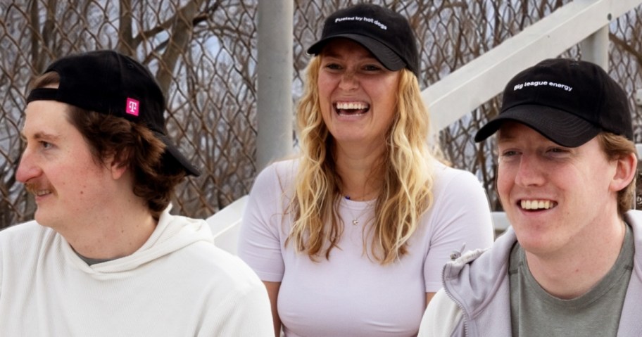 Three people wearing black t-mobile baseball caps are sitting outdoors, smiling and laughing. The background features a metal fence and trees.