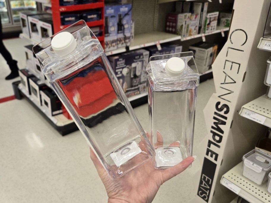 A hand holds two clear, empty rectangular bottles with white caps in a store aisle. The background features shelves of boxed kitchen items and storage containers.