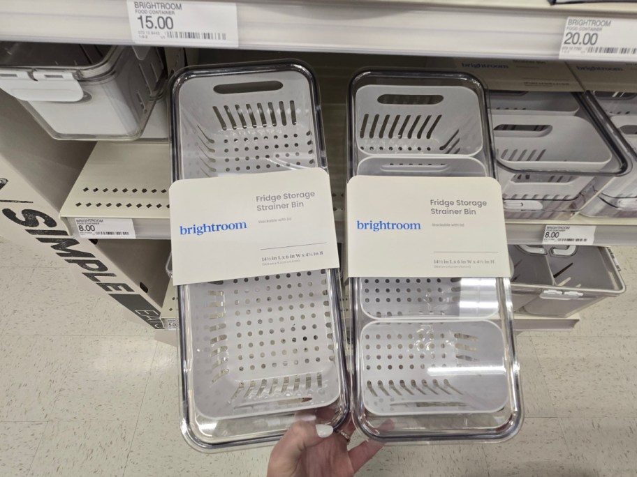 Hands holding two clear fridge storage strainer bins with labels, in a store aisle. Other bins are neatly arranged on shelves in the background.