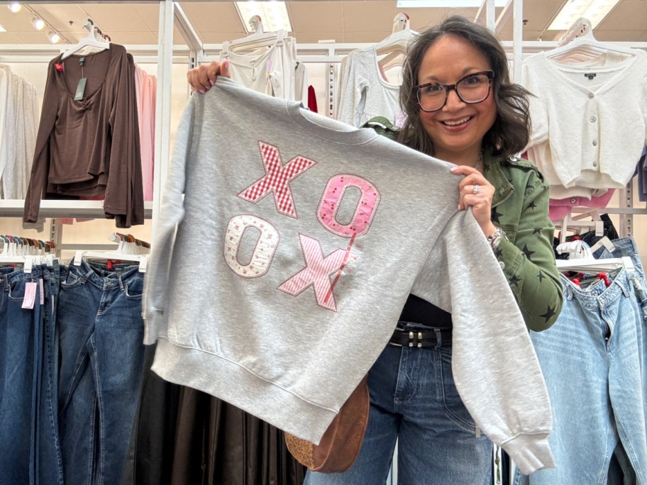 Woman smiling in a store, holding a gray sweatshirt with colorful "XOXO" appliqu&eacute;.