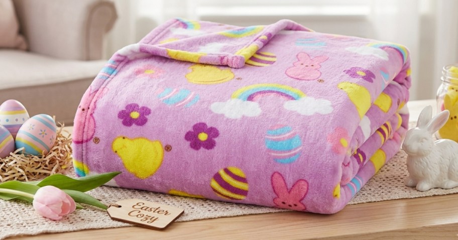 A plush, pink blanket with Easter-themed patterns featuring eggs, bunnies, and flowers, lies folded on a table. Nearby are painted eggs and a small rabbit figure.