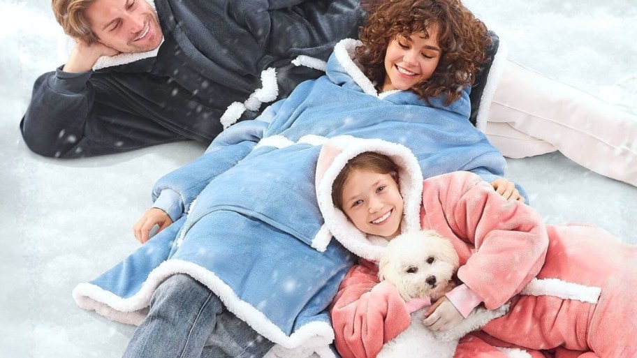 a dad, mom and daUGHTER WERAING HOODED BLANKETS AND LOUNGING WITH A PET DOG