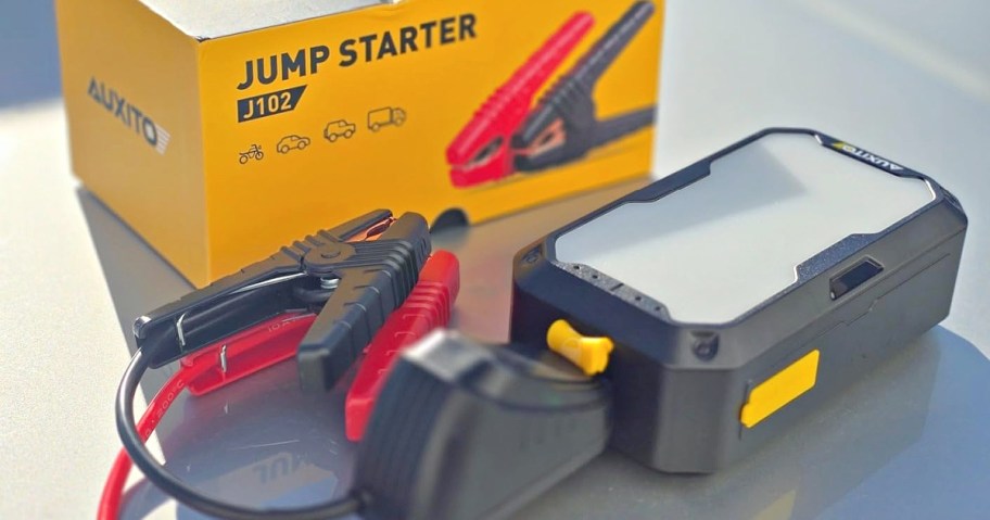 Portable jump starter kit with red and black clamps, yellow accents, and a compact black device. Background features a branded box and sunny lighting.