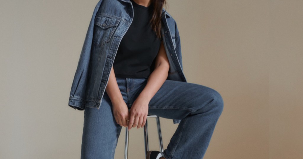 Women sitting on stool wearing black T-shirt and blue jeans with jean jacket draped on shoulders