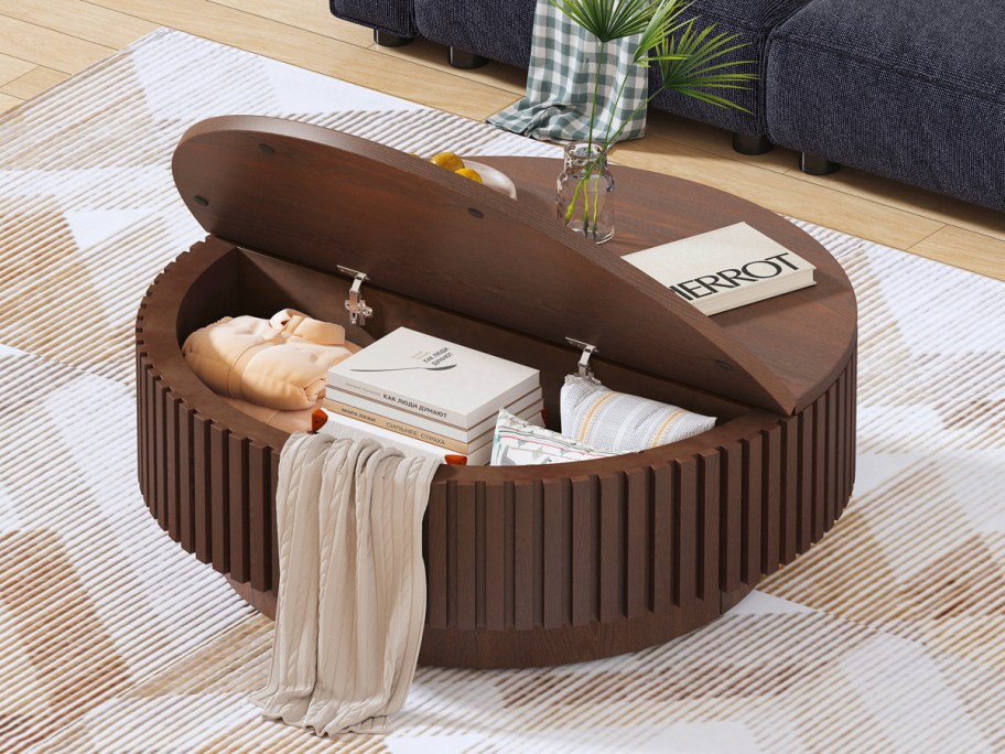 Round wooden coffee table with open lid revealing storage. Items include books, a folded blanket, and a vase on top.