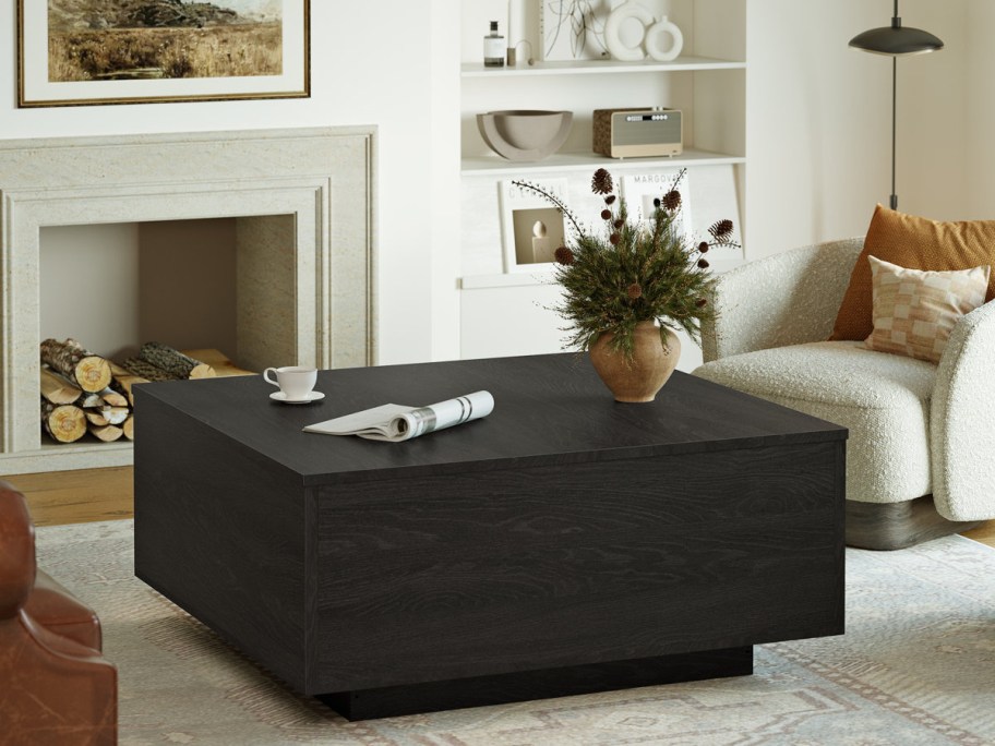 A cozy living room with a black coffee table holding a teacup, newspaper, and vase of flowers. A cushioned armchair sits nearby; a fireplace in the background.