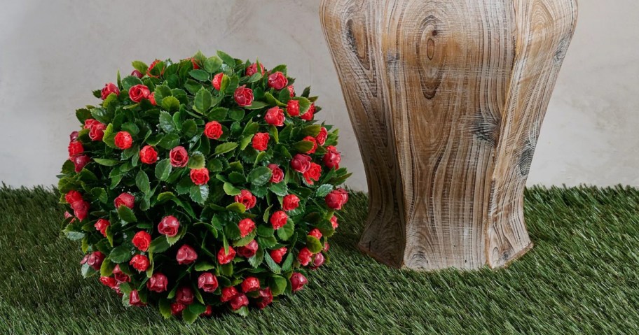 A spherical arrangement of red roses with green leaves sits on artificial grass next to a tall, wooden-textured vase.
