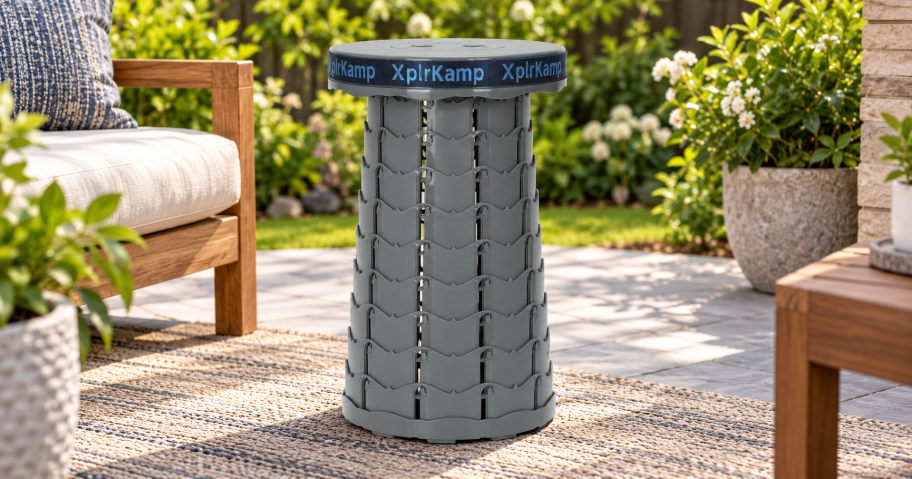 Gray XplrKamp collapsible stool fully extended on a patio beside outdoor seating and potted plants.