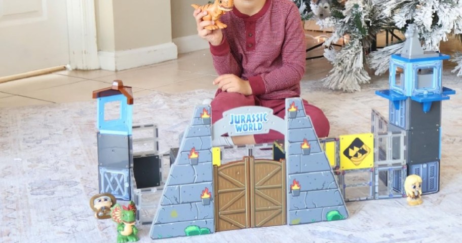 A child in a red outfit sits on a white carpet near a toy Jurassic World playset, holding a dinosaur figure. A decorated Christmas tree is in the background.
