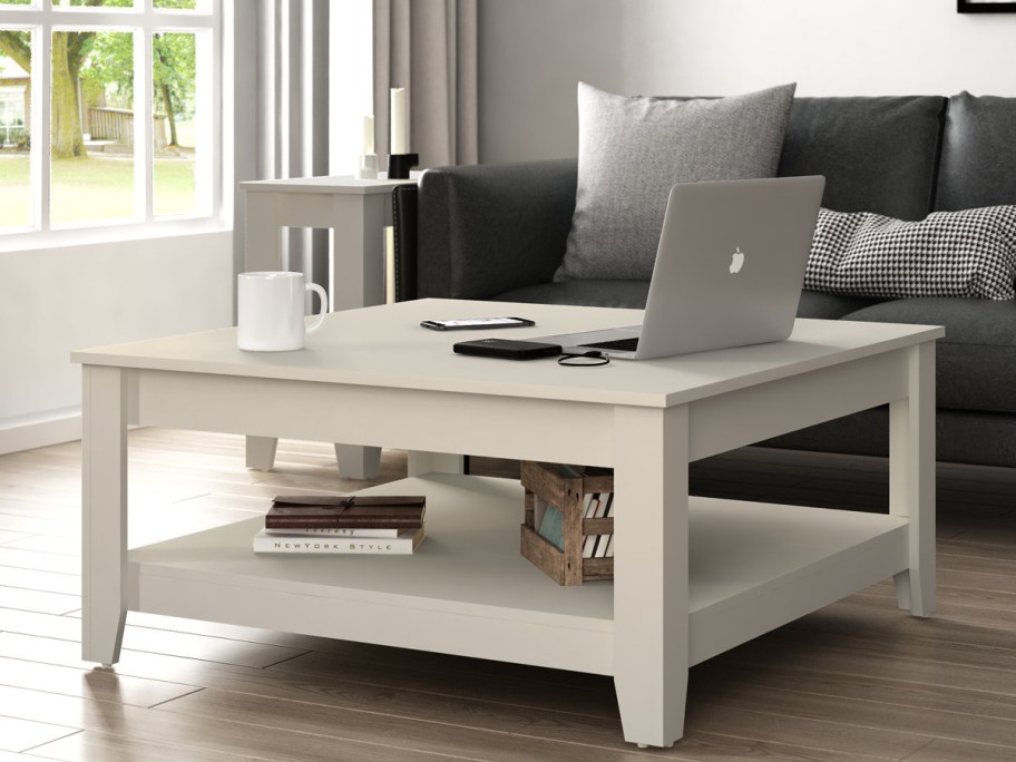 A cozy living room features a white coffee table with a laptop, phone, and mug. A dark gray sofa and window with trees are outside.