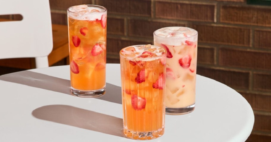 Three iced Starbucks beverages with strawberries sit on a white table, casting shadows. The drinks have a refreshing appearance, set against a brick wall backdrop.