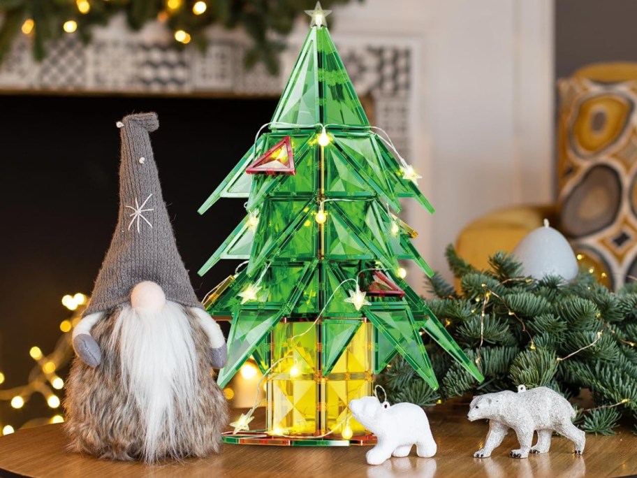 A green magnetic tile Christmas tree illuminated with string lights, displayed on a tabletop next to a plush gnome and polar bear figurines.