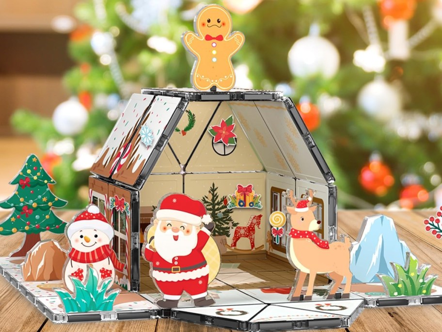 A PicassoTiles 45-piece magnetic gingerbread house building set with holiday decorations, a gingerbread man topper, and festive character figures displayed on a wooden surface in front of a Christmas tree.