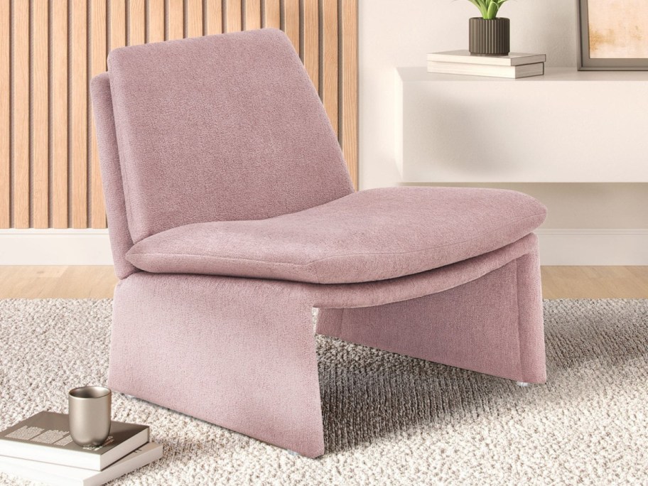 A mauve modern lounge chair sits on a textured rug in a cozy room. A white shelf holds books and a plant, with a warm wooden panel backdrop.