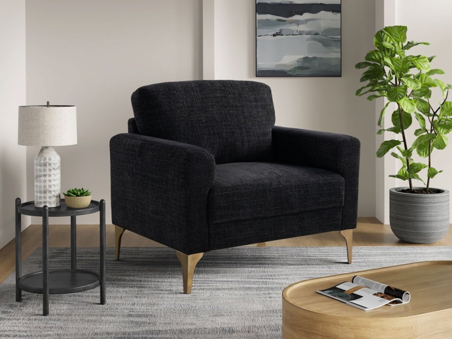 A modern living room features a dark fabric armchair with wooden legs, a round side table with a lamp and plant, and a large potted plant on a striped rug.