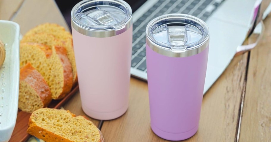 a pink and a purplish pink travel tumbler next to bread and a laptop