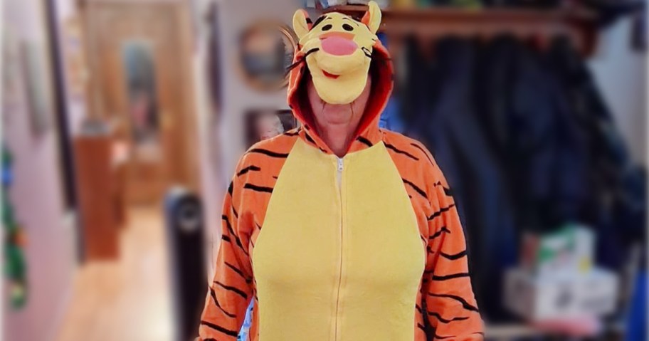 A person wearing a Tigger costume, with orange and black stripes, stands indoors. Their face is obscured by the hood.