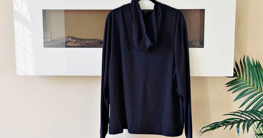 A black long-sleeve hoodie hangs on a white rectangular frame set against a beige wall. A green plant is partially visible on the right.