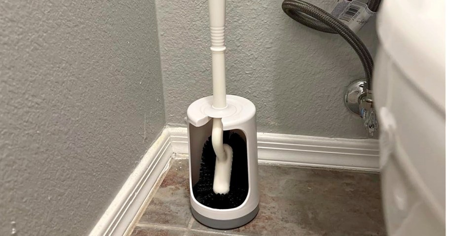 A white toilet brush with a black base sits in a holder beside a toilet on a tiled floor.