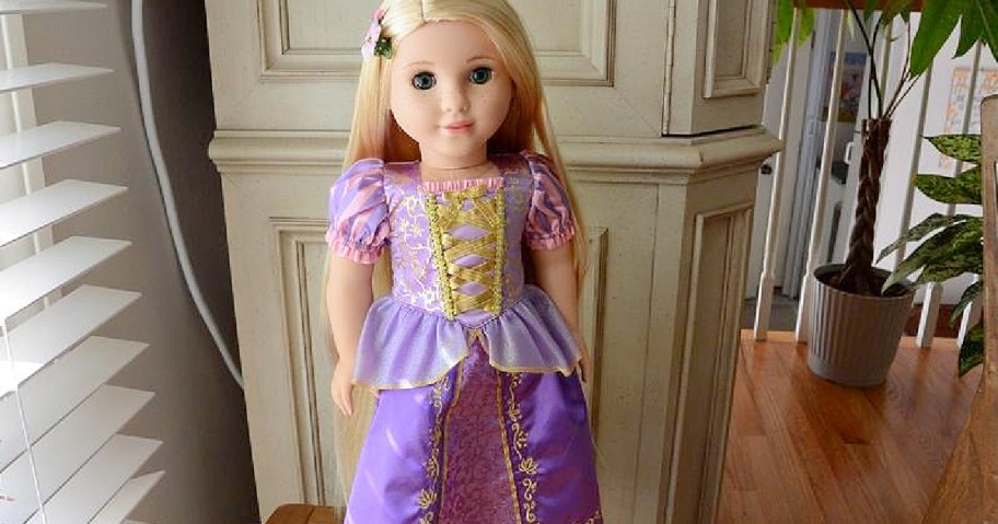 Blonde Rapunzel American Girl doll wearing a purple and gold dress, standing indoors near white shutters.