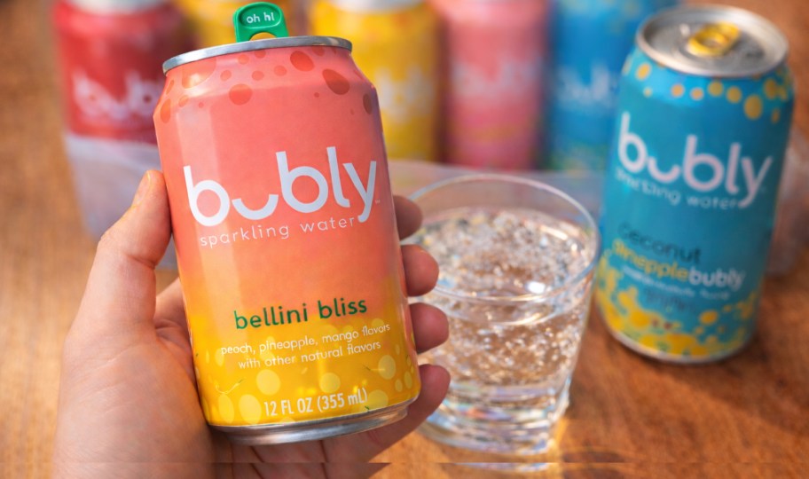 hand holding bubly sparkling water can with variety pack and glass on table