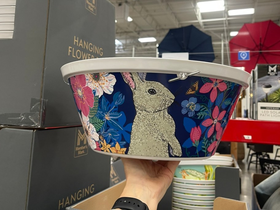 A hand holding a decorative hanging flower pot featuring a rabbit and colorful flowers, with other merchandise visible in the background.
