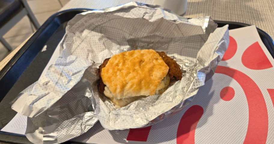 Chick fil is a chicken biscuit sandwich that sits in a wrapper on a tray.