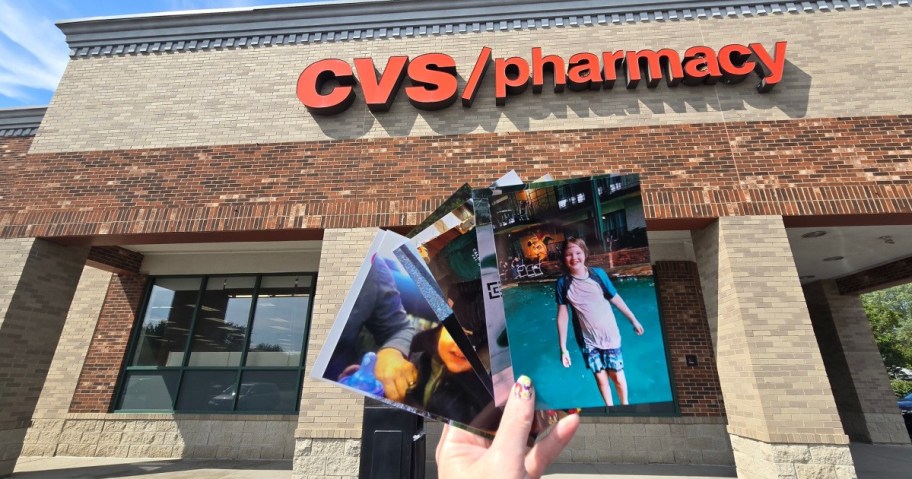 A hand holding printed photos of a child in front of a CVS/pharmacy store. The brick building and red signage are prominently visible.