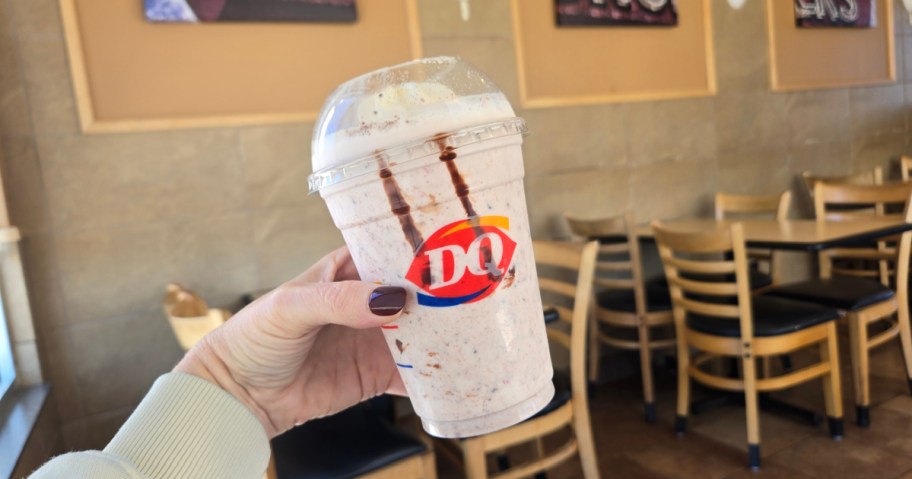 A hand holds a Dairy Queen milkshake with whipped cream in a clear cup inside a cozy restaurant. Empty wooden tables and chairs are in the background.