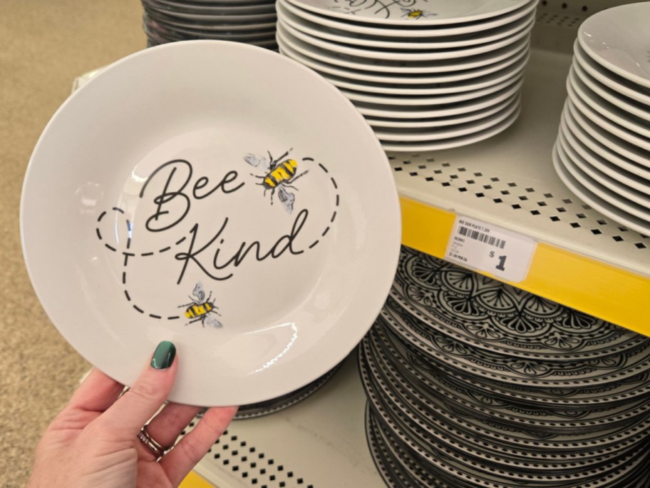 A hand holds a white plate with "Bee Kind" text and bee illustrations. More plates are stacked on shelves in the background, priced at $1.