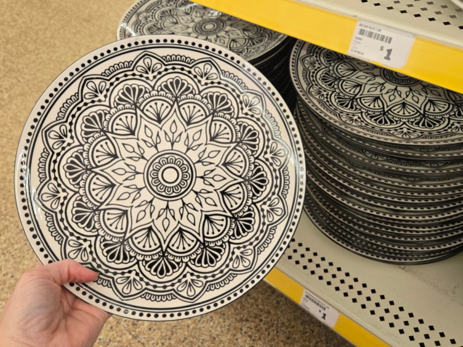 A hand holds a white plate with intricate black mandala patterns. Nearby, similar plates are stacked on a store shelf, priced at $1 each.