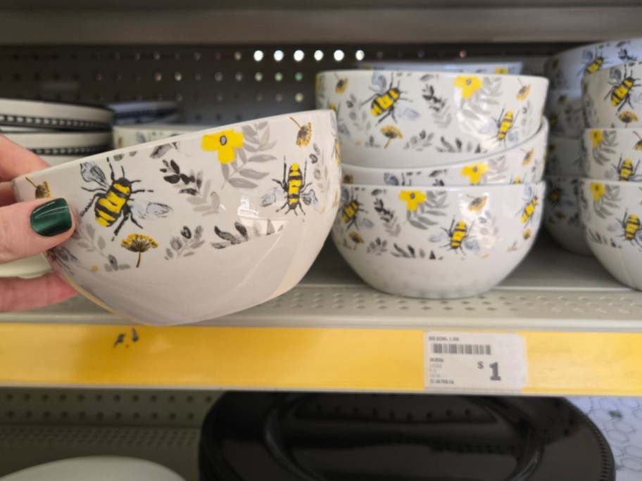 A person holds a ceramic bowl with a bee and floral pattern. Several similar bowls are displayed on a store shelf. Price tag reads $1.