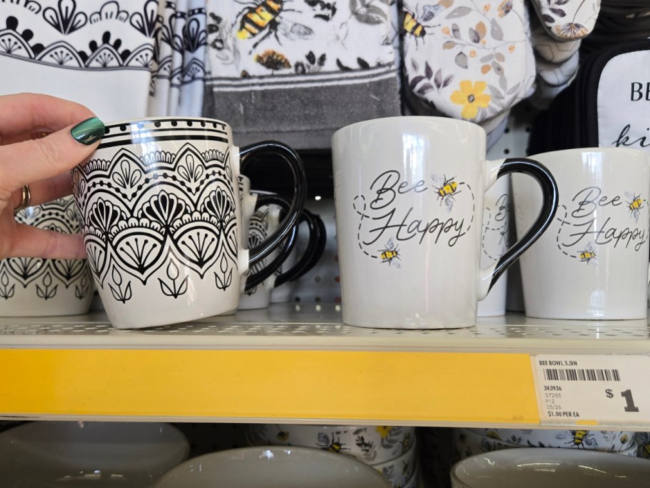 A hand holds a white mug with black geometric patterns next to a mug that reads "Bee Happy" with bee illustrations. Both are on a store shelf.