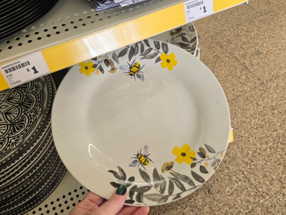 A white ceramic plate with yellow flowers and bee designs is shown in a store display. It is priced at $1, set against a wooden background.