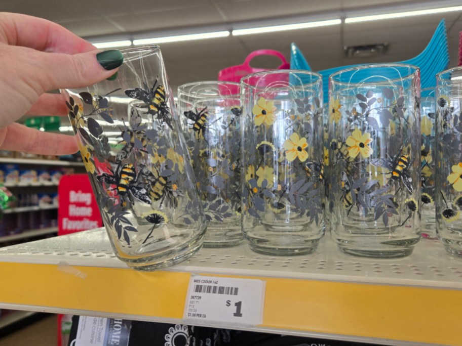Hand holding a glass with bee and flower pattern, on display with similar glasses on a store shelf. Shelf tag shows the price as $1.