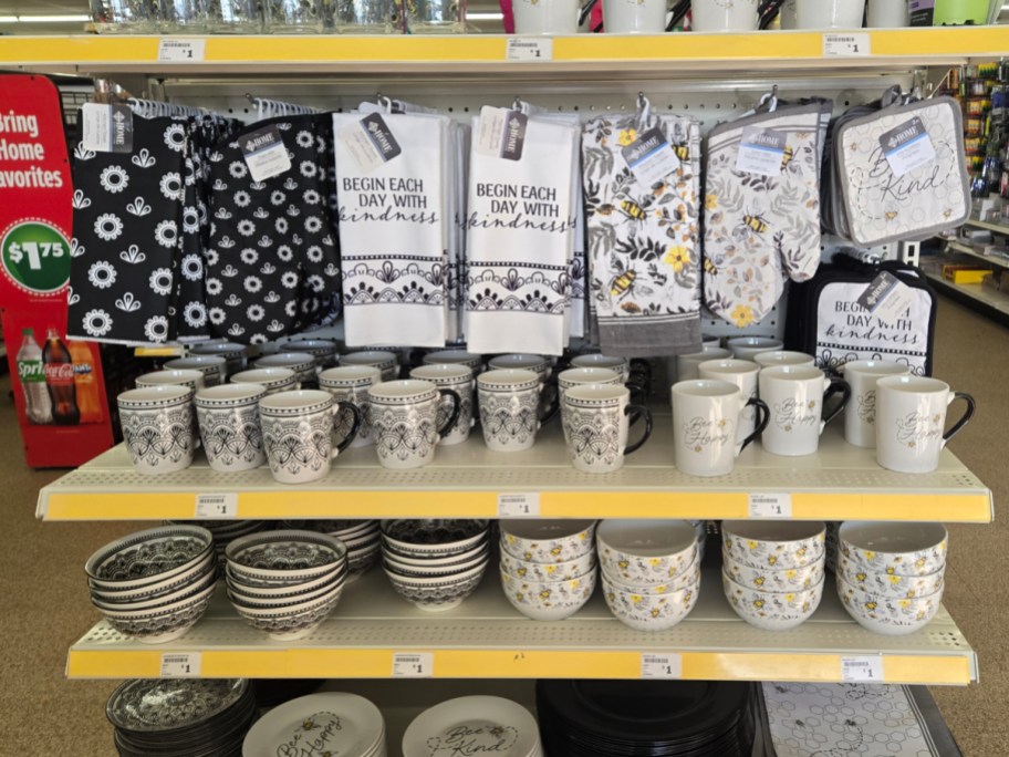 Store display with black and white kitchenware: mugs, bowls, and towels with floral patterns and "Begin Each Day with Kindness" text.