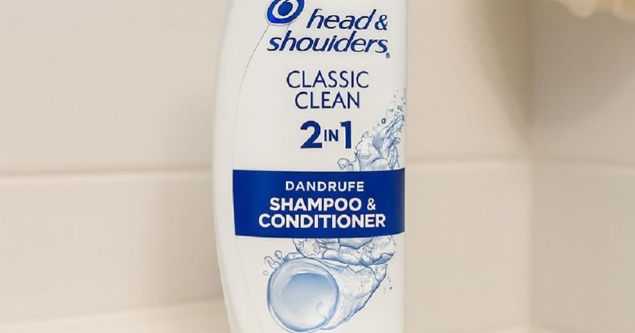 Head & Shoulders Classic Clean 2 in 1 shampoo and conditioner bottle on a bathroom shelf. White bottle with blue text and water splash graphic.