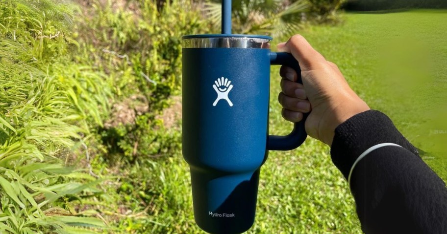 A person holding a blue Hydro Flask tumbler with a straw in an outdoor setting with greenery in the background.