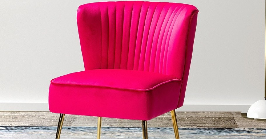 A vibrant pink velvet chair with a ribbed backrest and gold legs is set on a wooden floor.