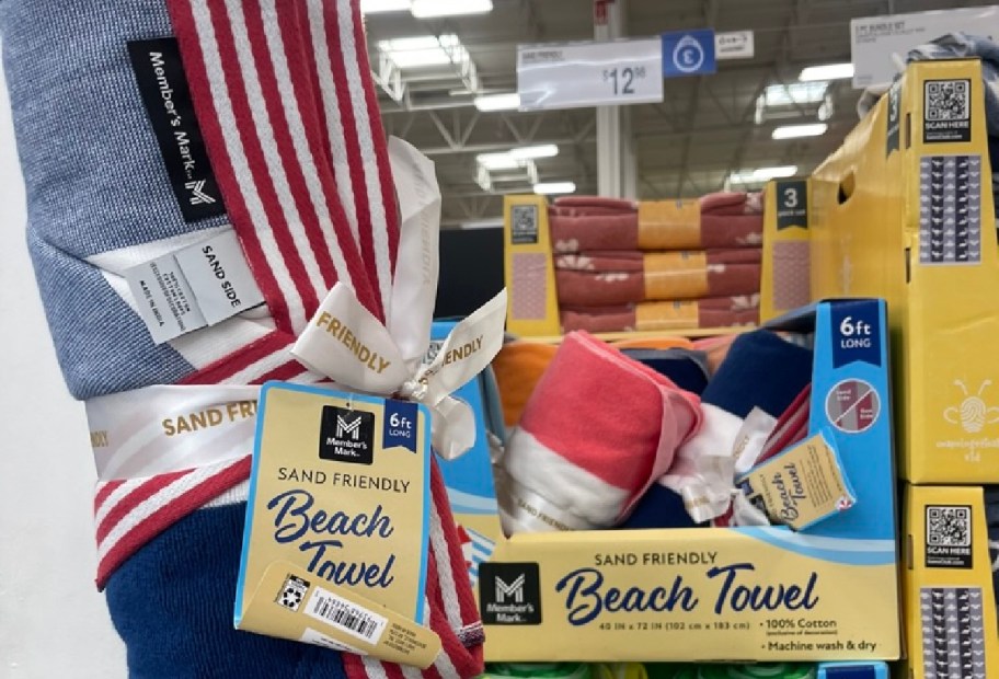 A display of beach towels, including a blue towel with a red and white striped design, wrapped with a ribbon that reads "Sand Friendly Beach Towel," along with other towels in various colors.
